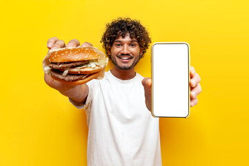 hungry Hindu guy holding tasty big burger and showing blank smartphone screen on yellow isolated...