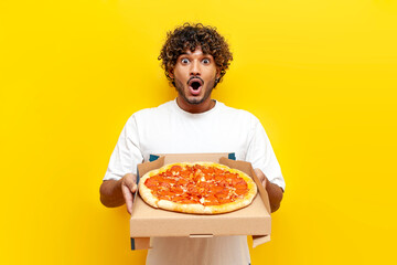hungry hindu guy holding a box with delicious pizza and surprised on yellow isolated background