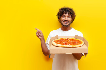 hungry guy indian holds box with delicious pizza and points with his hand at copy space on yellow...