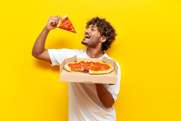 hungry guy indian holds box with delicious pizza and bites a piece on yellow isolated background