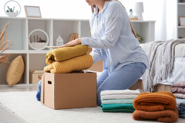 Young woman packing stack of clothes into cardboard box in bedroom - obrazy, fototapety, plakaty