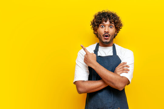 young surprised guy indian waiter in apron pointing at copy space on yellow isolated background