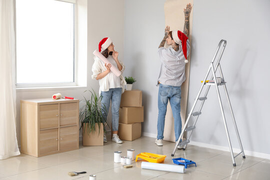 Young pregnant couple with Santa hats wallpapering during repair in their new house