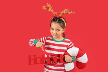 Little girl in reindeer horns holding Christmas cushion and pointing at word HOHOHO on red background
