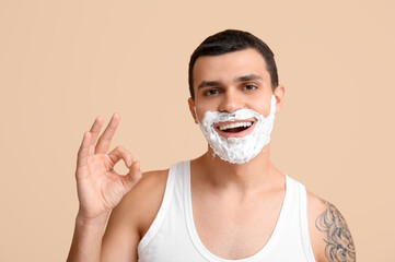 Handsome young man with shaving foam on his face showing OK gesture against beige background