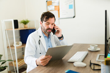Handsome latin doctor talking on the phone with a patient while looking at the medical record working at his office