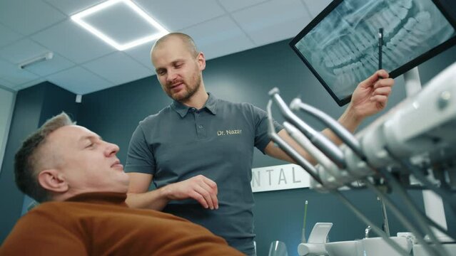 Smiling dentist pointing on screen with MRI scan of teeth, giving consultation to man having dental problems, sitting in chair in modern dentist office. High quality 4k footage