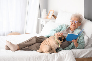 Senior woman with cute pug dog reading book in bedroom