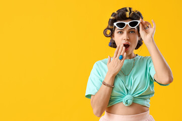 Shocked young woman with hair curlers on yellow background