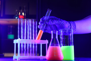 Scientist working with laboratory glassware of luminous liquids at table against dark background,...