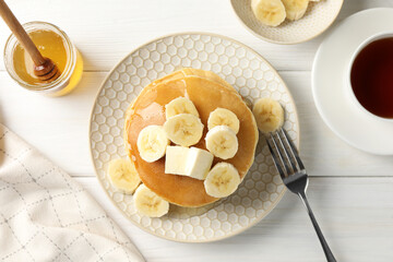 Delicious pancakes with bananas, honey and butter served with tea on white wooden table, flat lay
