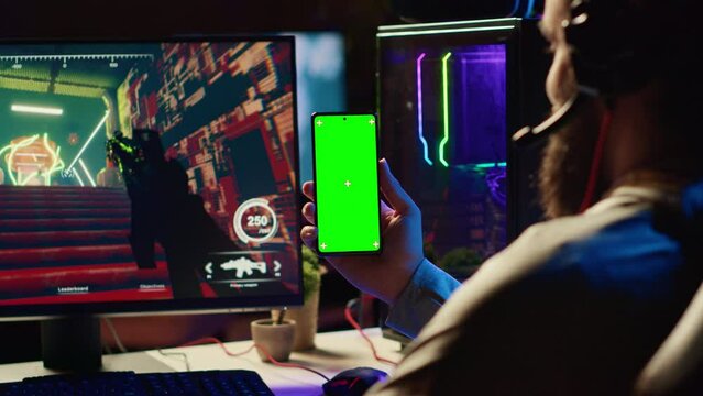 Man watching tutorial on green screen phone while playing first person shooter videogame with gun shooting laser bullets. Gamer learning to play game by looking at online guide on smartphone