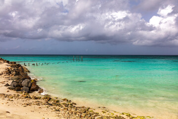 Tropical beach on the island of Aruba. Sandy beach in foreground. Emeral water with remains of old pier stretching from shore. Cloudy sky in the distance. 
