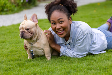 African American woman is playing with her french bulldog puppy while lying down in the grass lawn after having morning exercise in public park