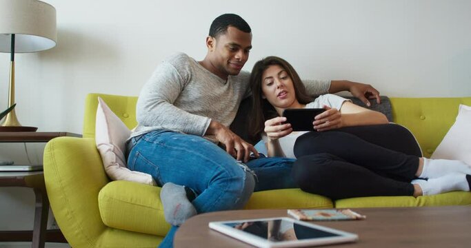 Young millennial couple watching video on phone as they sit on the couch. African American and Caucasian boyfriend and girlfriend in their 20s hanging out in their apartment. 4k Slow motion handheld