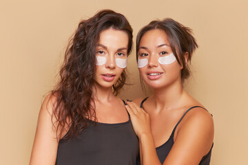 Two pretty girlfriends stand on studio background close to each other both have patches under eyes, skincare concept, copy space