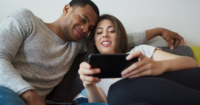 Young millennial couple laughing at a cute pictures on social media using phone. African American and Caucasian boyfriend and girlfriend watching funny videos online. 4k Slow motion handheld