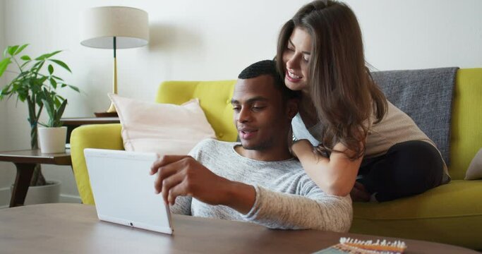 Happy young millennial couple using tablet computer in living room. African American and Caucasian boyfriend and girlfriend in apartment watching video on pad. 4k Slow Motion Handheld