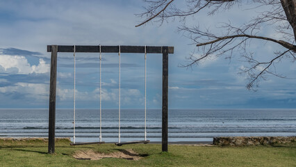 A place of rest by the sea. Two wooden swings are suspended from a crossbar. Ahead is the turquoise ocean. The waves are rolling towards the shore. Tree branches against a  blue sky and clouds. 