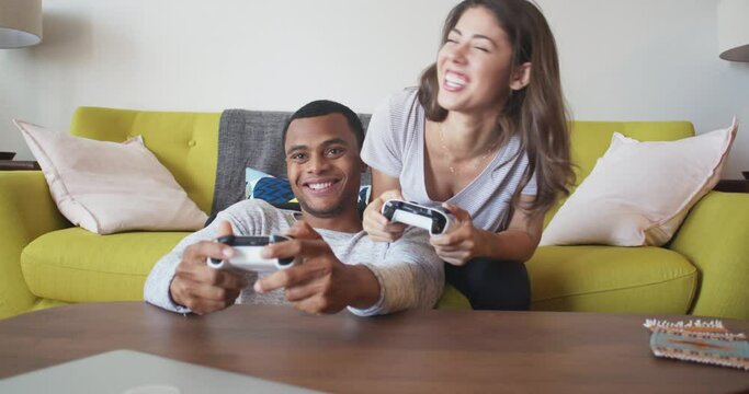 Happy young millennial couple gaming in living room. Competitive and playful African American and Caucasian boyfriend and girlfriend playing multiplayer console video game. 4k slow motion handheld