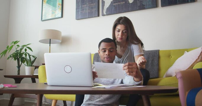 Young African American and Caucasian Millennial couple paying bills online in their living room. New home owners going over papers together. 4k Slow motion handheld