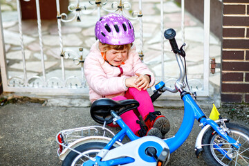 Cute little girl sitting on the ground after falling off her bike. Upset crying preschool child with safe helmet getting hurt while riding a bicycle. Active family leisure with kids.