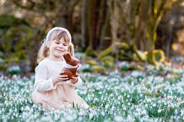 Happy little girl with Easter bunny ears eating chocolate figure in spring forest on sunny day,...