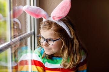 Portrait of a happy little girl with bunny ears looking outside sitting by a window. Easter holiday. Adorable child happy about holiday.