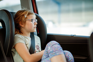 Little Preschool Girl Sitting in Her Car Seat. Happy Child with Eyeglasses looking out of the window. Bored kid on the Way to Family Vacations during Traffic Jam.