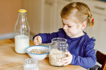 Adorable toddler girl eating healthy oatmeals with milk for breakfast. Cute happy baby child in...