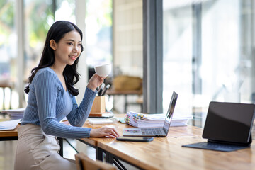 Beautiful young smiling Asian businesswoman working on laptop and drinking coffee, Asia businesswoman working document finance and calculator in her office.
 - obrazy, fototapety, plakaty