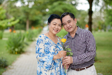 Fototapeta premium Portrait of happy senior Vietnamese couple hugging and holding rose flower when standing on path in city park