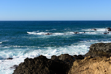 Turbulent ocean waves crashing against the rocky, rugged shoreline under clear blue sky of Russian...