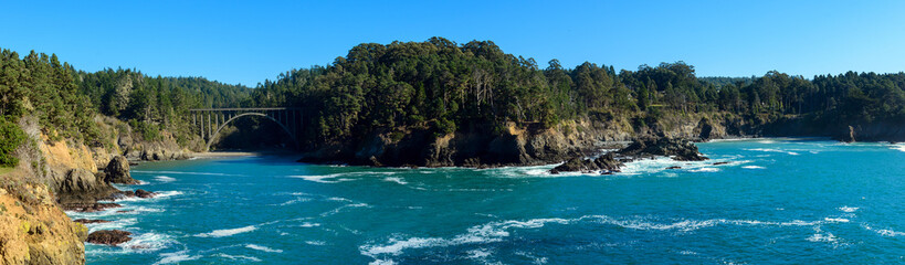 Panoramic scenic view of of azure ocean waters bordered by rocky cliffs and verdant forests near Russian Gulch Bridge