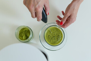Woman is mixing matcha powder with hot water in glass cup by electric whisk 