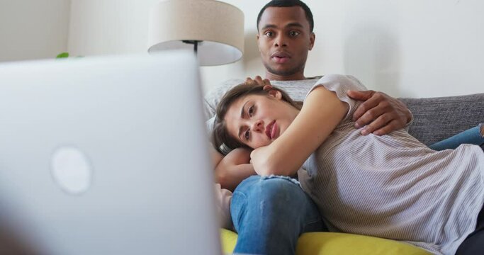 Millennial couple watching scary movie online with their laptop. African American and Caucasian boyfriend and girlfriend scared by horror film streaming on computer. 4k slow motion handheld