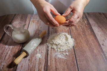 woman breaking an egg, pouring it over flour to make a sponge cake on a wooden table with a rolling...