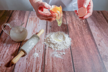woman breaking an egg, pouring it over flour to make a sponge cake on a wooden table with a rolling pin and a pitcher of milk
