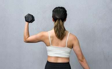 An athletic woman firmly grasps a hefty dumbbell behind her back, showcasing strength and determination. She stands in a well-lit gym atmosphere, the picture of healthful energy and dedication.