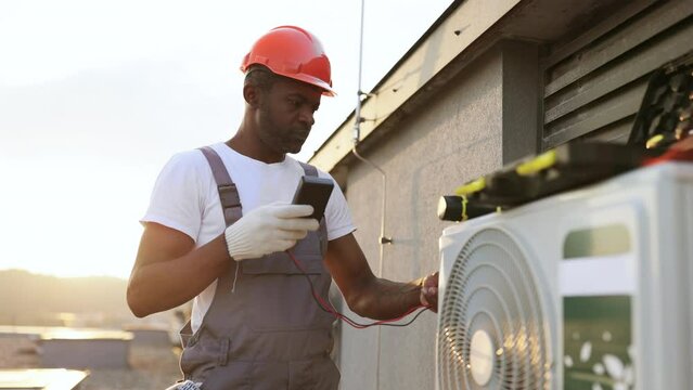 Focused african american specialist using voltage tester while installing modern cooling system on rooftop of residential building. Manager measuring current strength in hanging air conditioner.