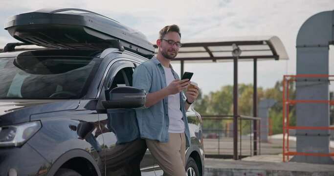 Young Caucasian man standing by the car with roof racks and cargo box and drinking coffee on a parking