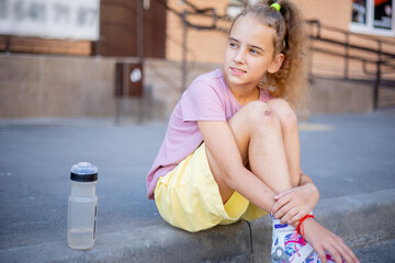 A pretty teenager is sitting on the sidewalk near a high-rise building and resting after a fun...