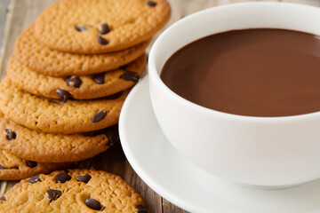 sweet snack. on a dark wooden table there is a white mug of cocoa on a saucer, next to there is a stack of cookies with chocolate
