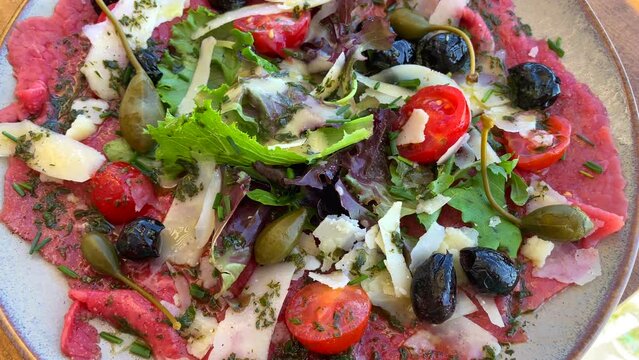 Delicious traditional gourmet beef carpaccio with parmesan cheese, cherry tomatoes, salad, capres, black olives and pesto sauce on a plate at a French Italian restaurant, 4K shot