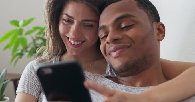 Young millennial couple watching a cute video on girlfriends cell phone while resting on the couch. African American and Caucasian man and woman using smartphone together. 4k slow motion handheld