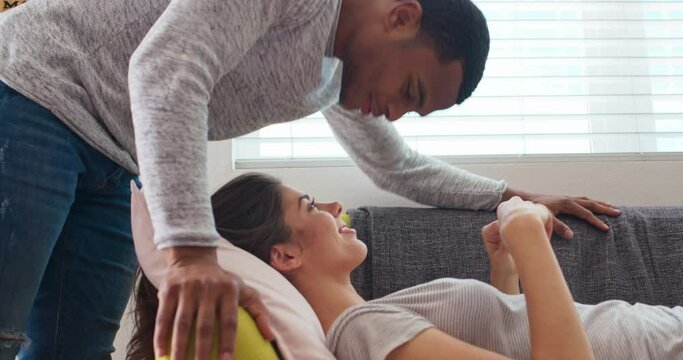 Attractive young black man entering frame and kissing his girlfriend as she lays on the couch. Caucasian and African American millennial couple share kiss in their living room. 4k slow motion handheld