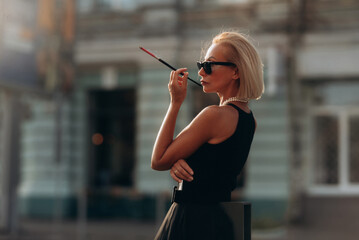 Senior woman smoking a cigarette outdoor. Stylish blonde woman in black sunglasses with mouthpiece - obrazy, fototapety, plakaty
