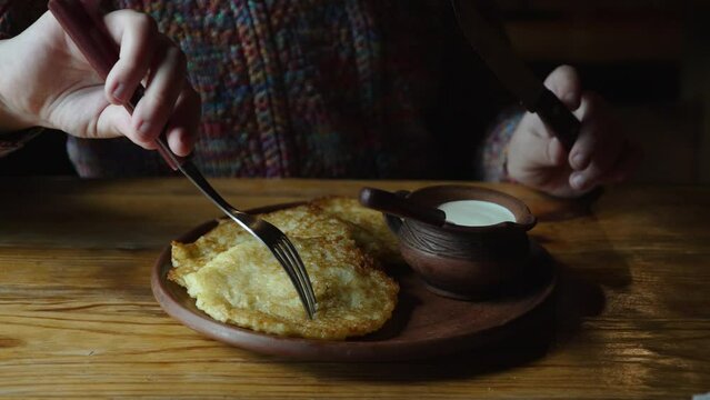 Left-handed boy with cutlery in hands cuts off piece of fried potato pancake. High calorie lunch for child in ethnic restaurant with dish in butter with sour cream. Closeup of served dish.
