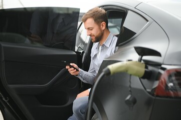 Happy man using smart phone and charging car at vehicle charging station
