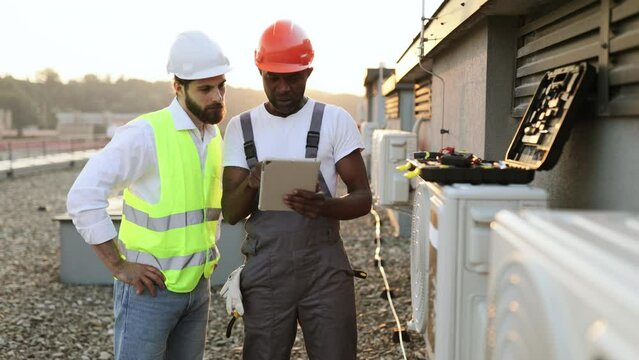 Multicultural males advising with each other about repairing of air conditioner while standing near broken cooling system outdoors. Men using tablet for scrolling websites with useful information.